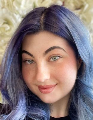 Portrait of a person with wavy blue hair, soft smile, and light makeup against a blurred cream background