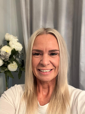 Smiling woman in white top before gray curtains, with white flowers to the left.