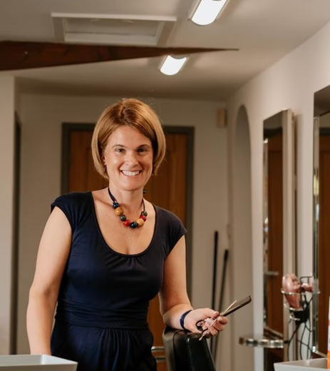 Woman standing in a salon holding a curling iron, smiling near mirrors and styling stations