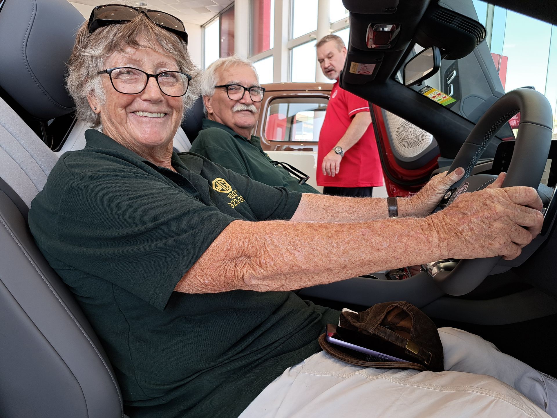 Woman smiles while sitting in a car's driver seat. Man in passenger seat smiles. Third man stands behind them.