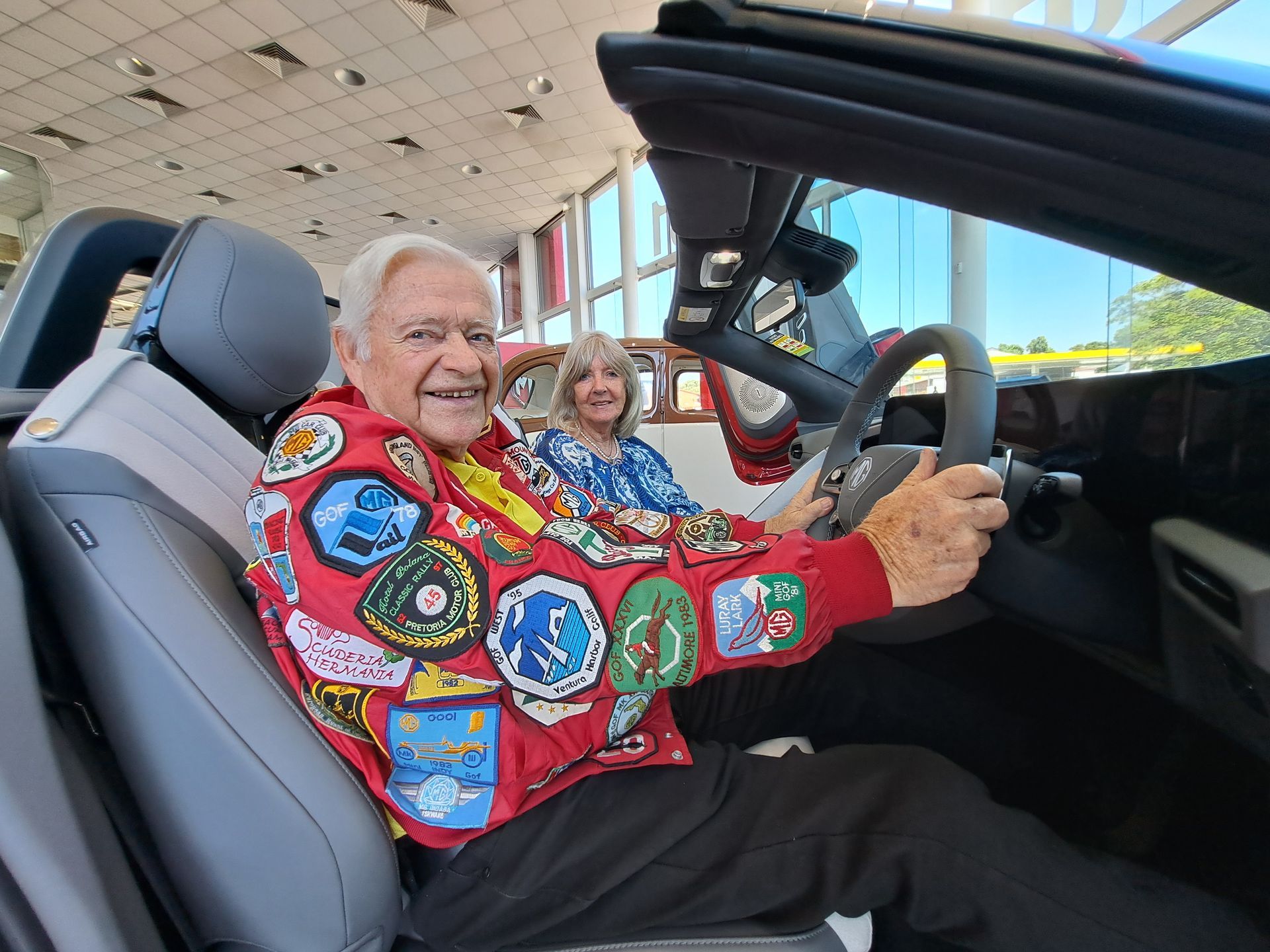 Man in a red jacket, smiling, sitting in a convertible car, woman in background, dealership.