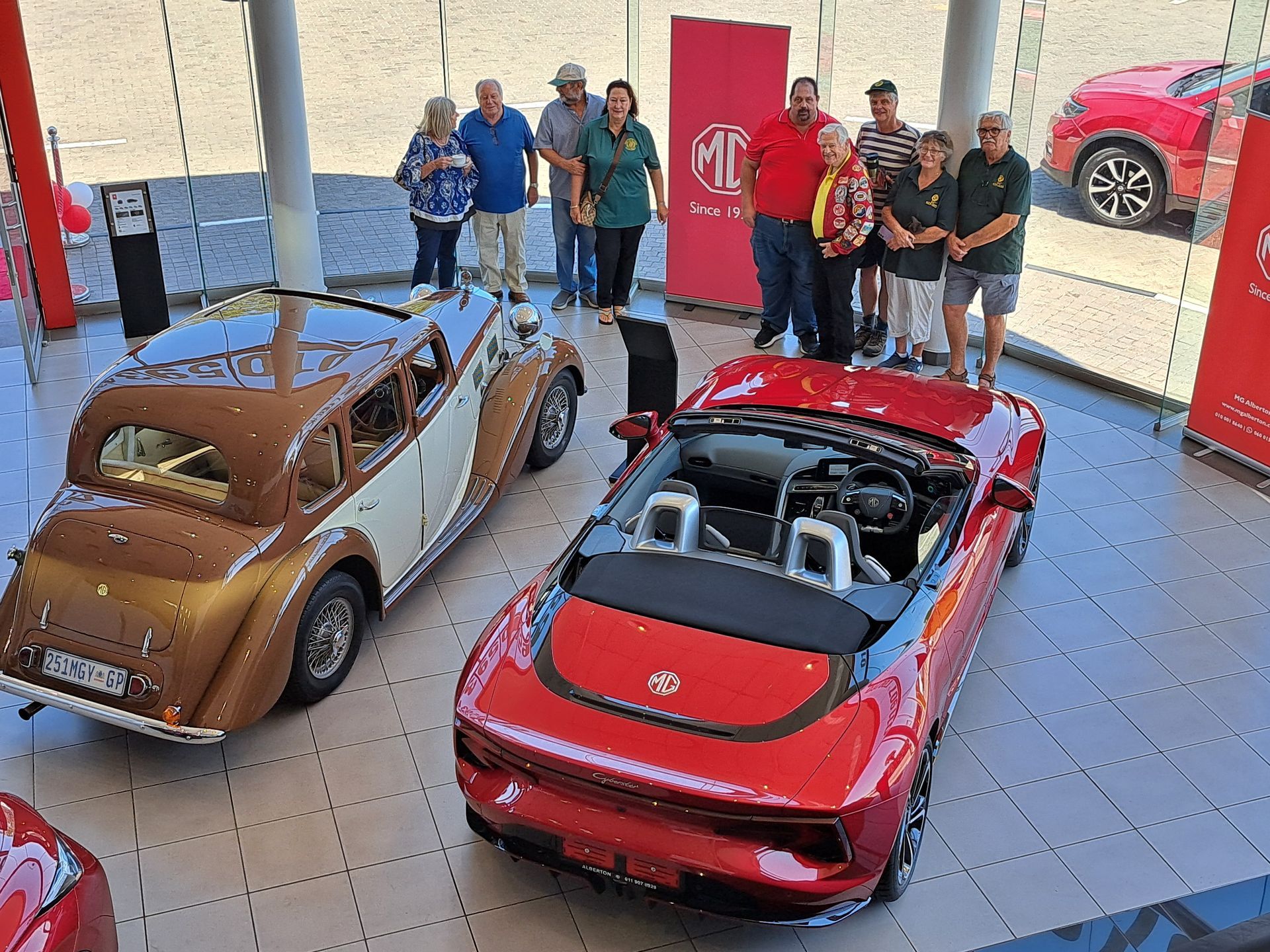 Two MG cars (brown classic & red convertible) displayed in a showroom with a group of people standing behind them.
