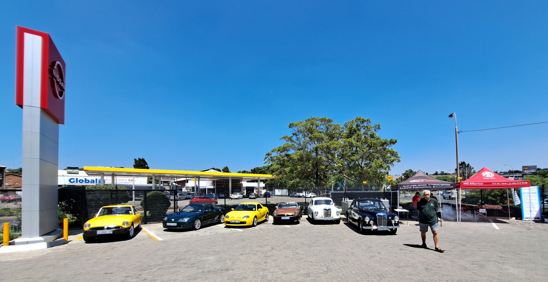 Classic cars parked near a gas station under a blue sky. A person stands nearby.
