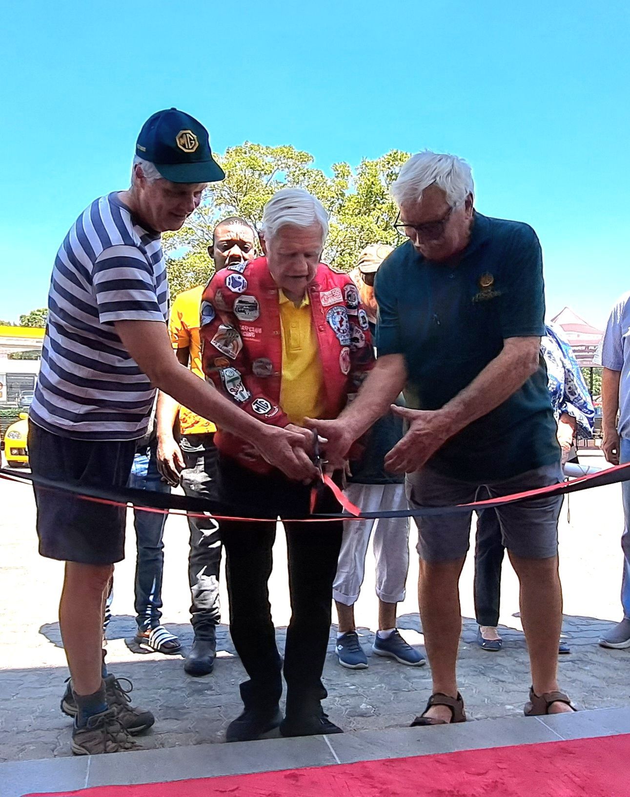 Three people cutting a red ribbon at an outdoor event. One wears a blazer, two have their hands on the scissors.