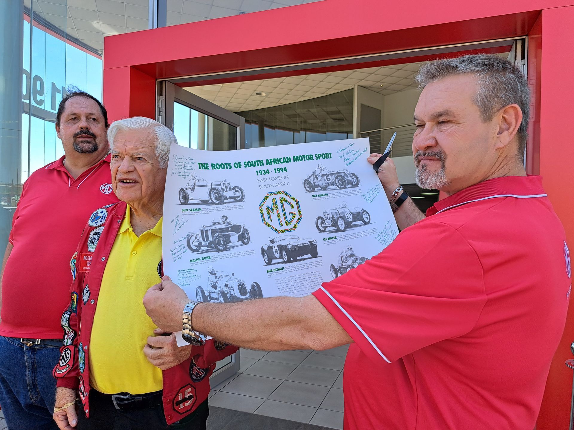 Three men holding a poster with MG car drawings. Exterior with red doorframe. One man in yellow shirt.