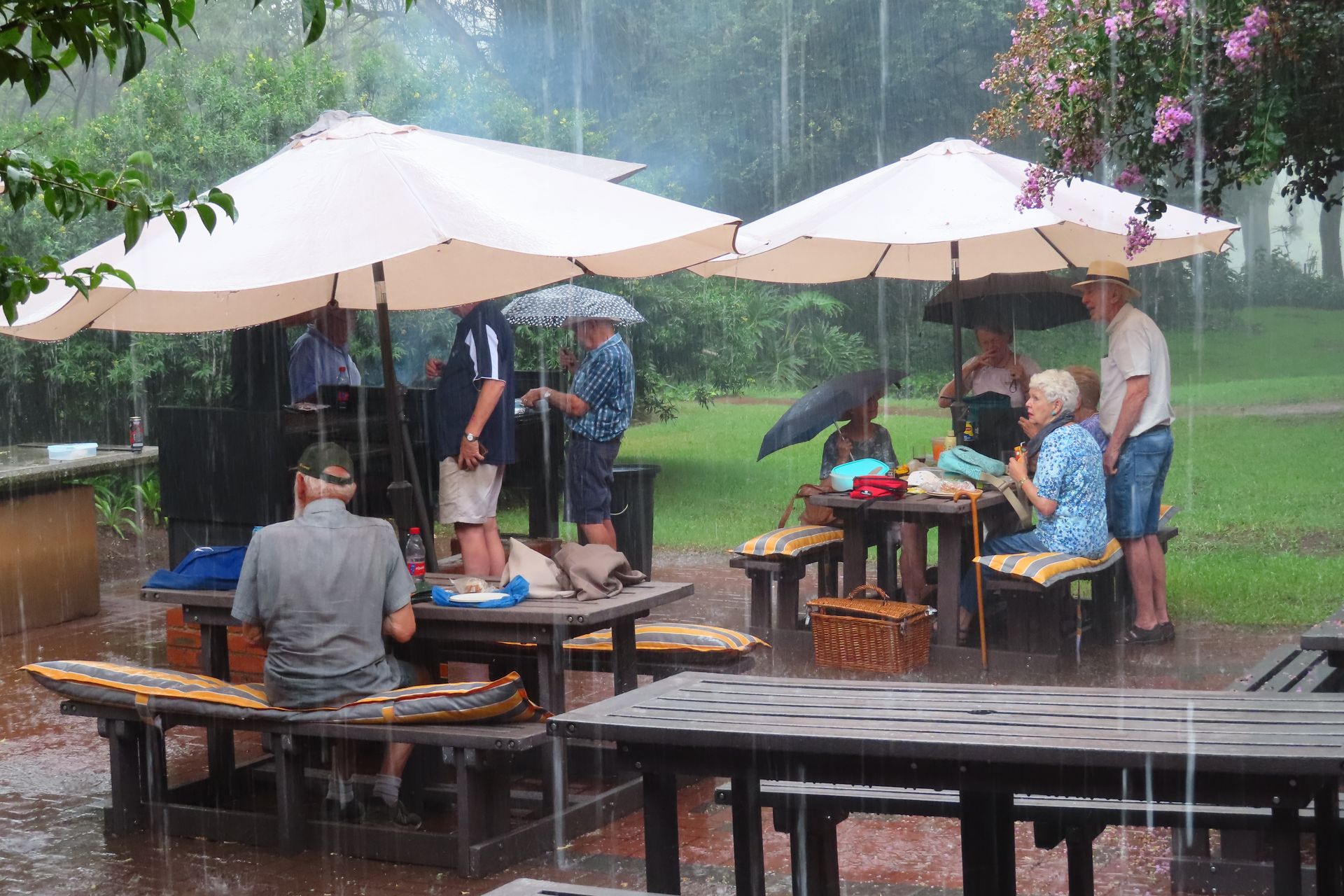 People gather outdoors under umbrellas and picnic tables in heavy rain.