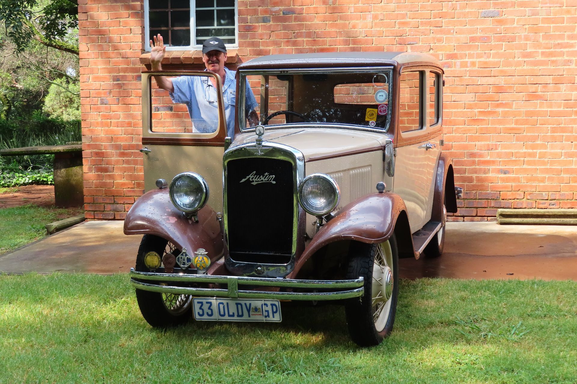 Man in a light blue shirt waving from a vintage tan and brown car parked on grass in front of a brick building.