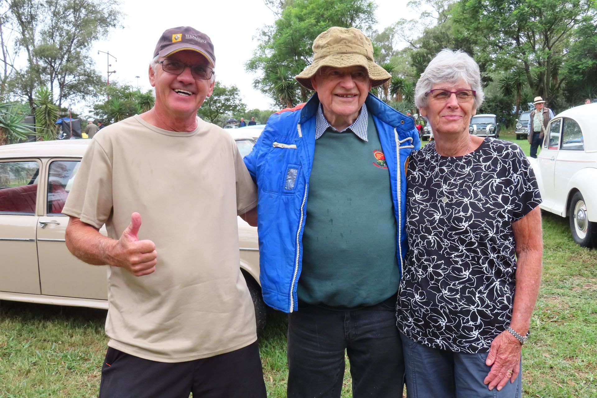 Three people smiling, posing together outdoors. Man in tan shirt gives thumbs up.
