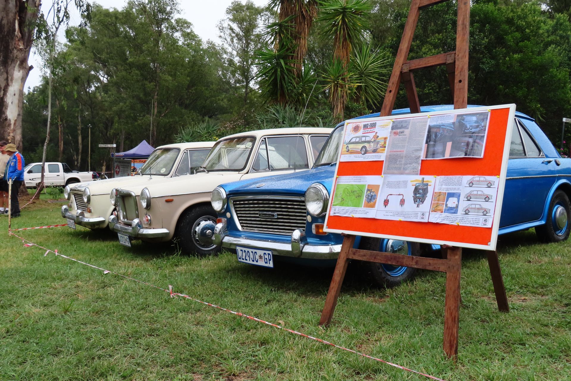 Classic cars on display at an outdoor event, blue and cream, with an easel holding information in front.