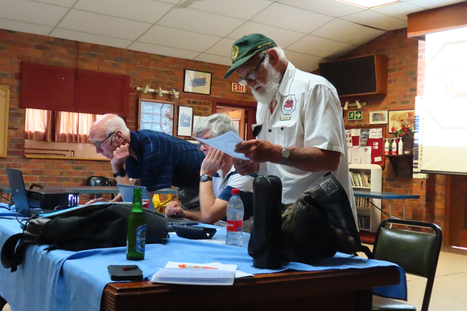 Men looking at papers, one wearing a hat, at a table with laptops, inside a brick-walled room.