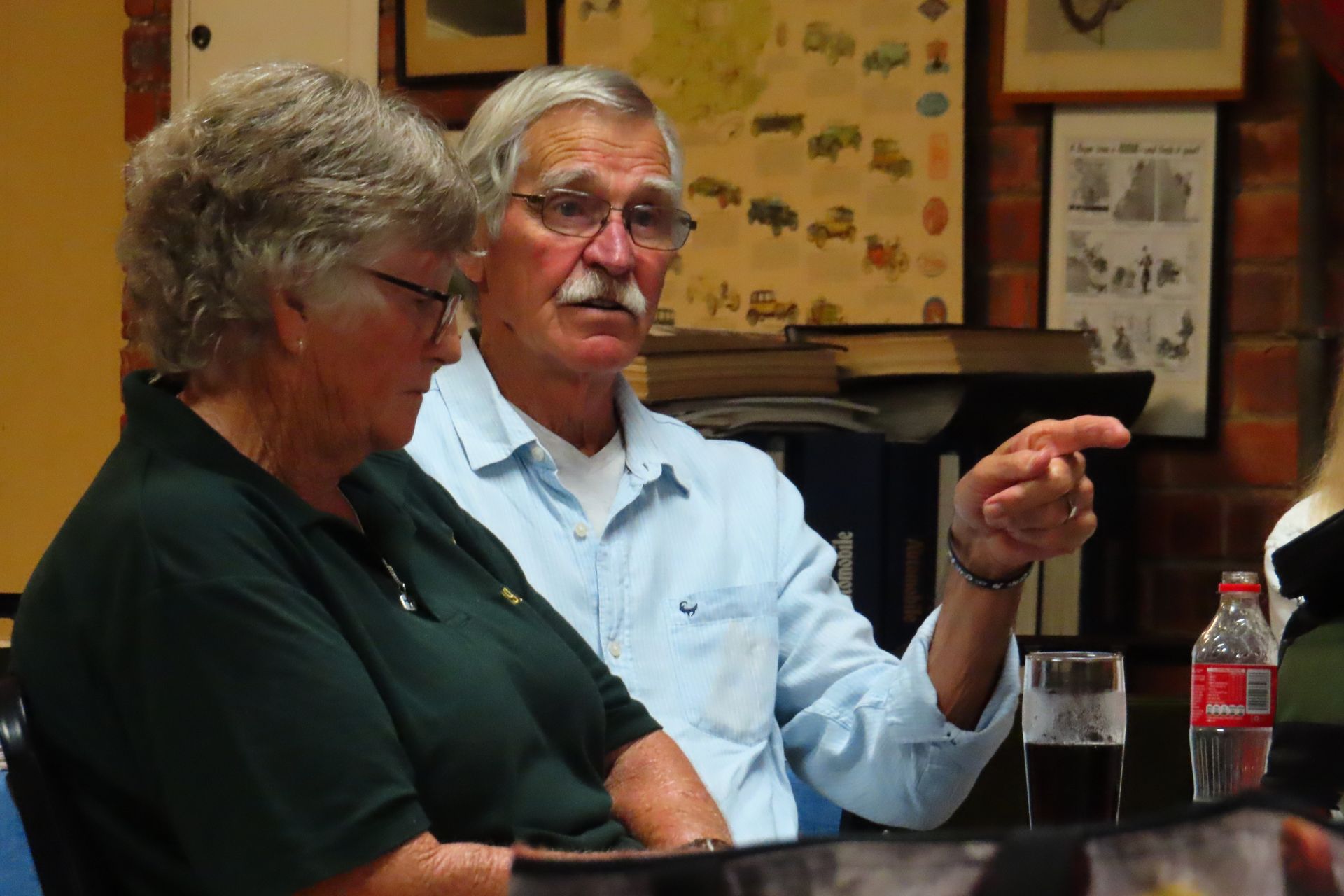 Man pointing, speaking to woman at a table. Bookshelves and wall art visible.