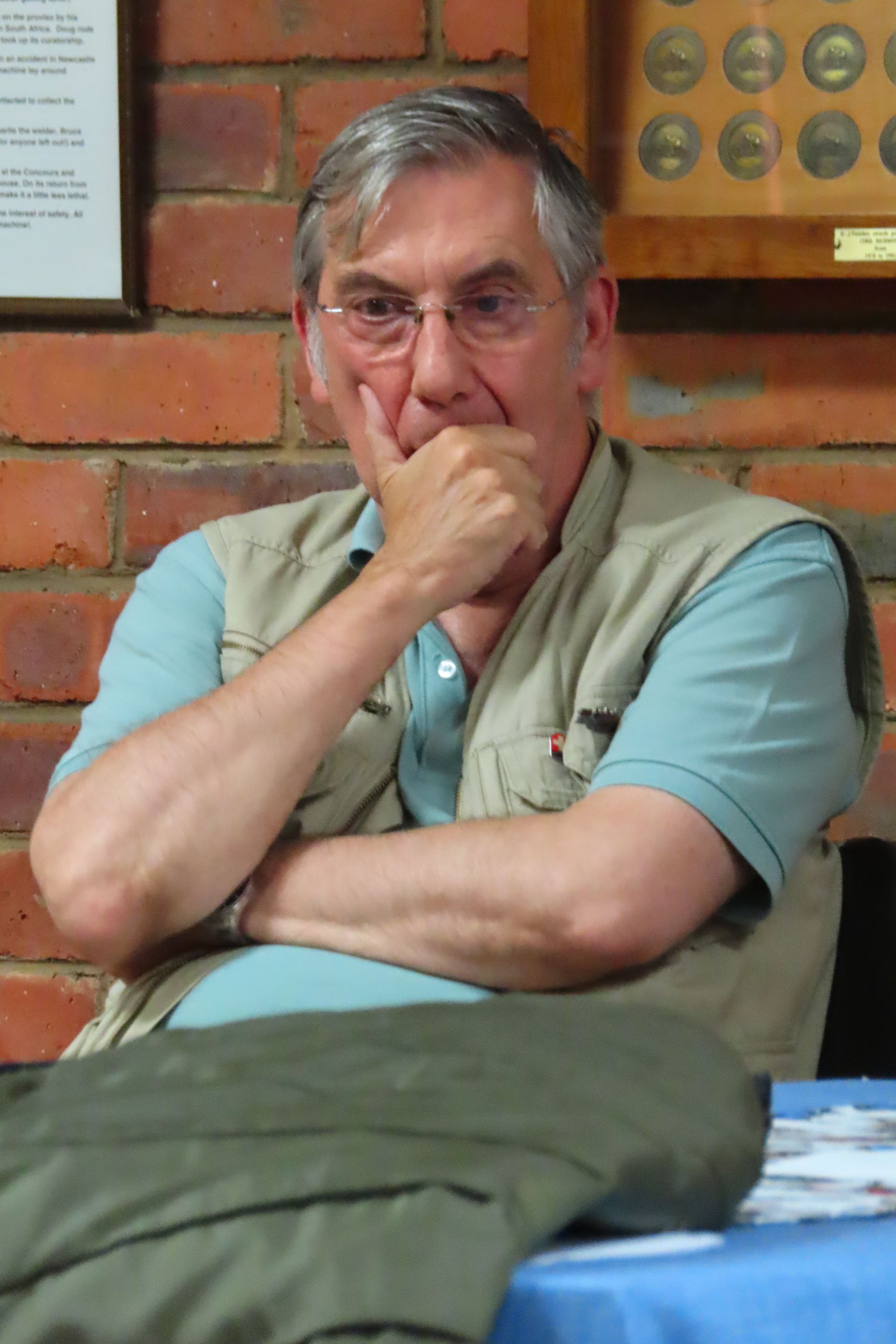 Man in glasses, resting chin on hand, wearing a light blue shirt and beige vest, seated indoors near a brick wall.