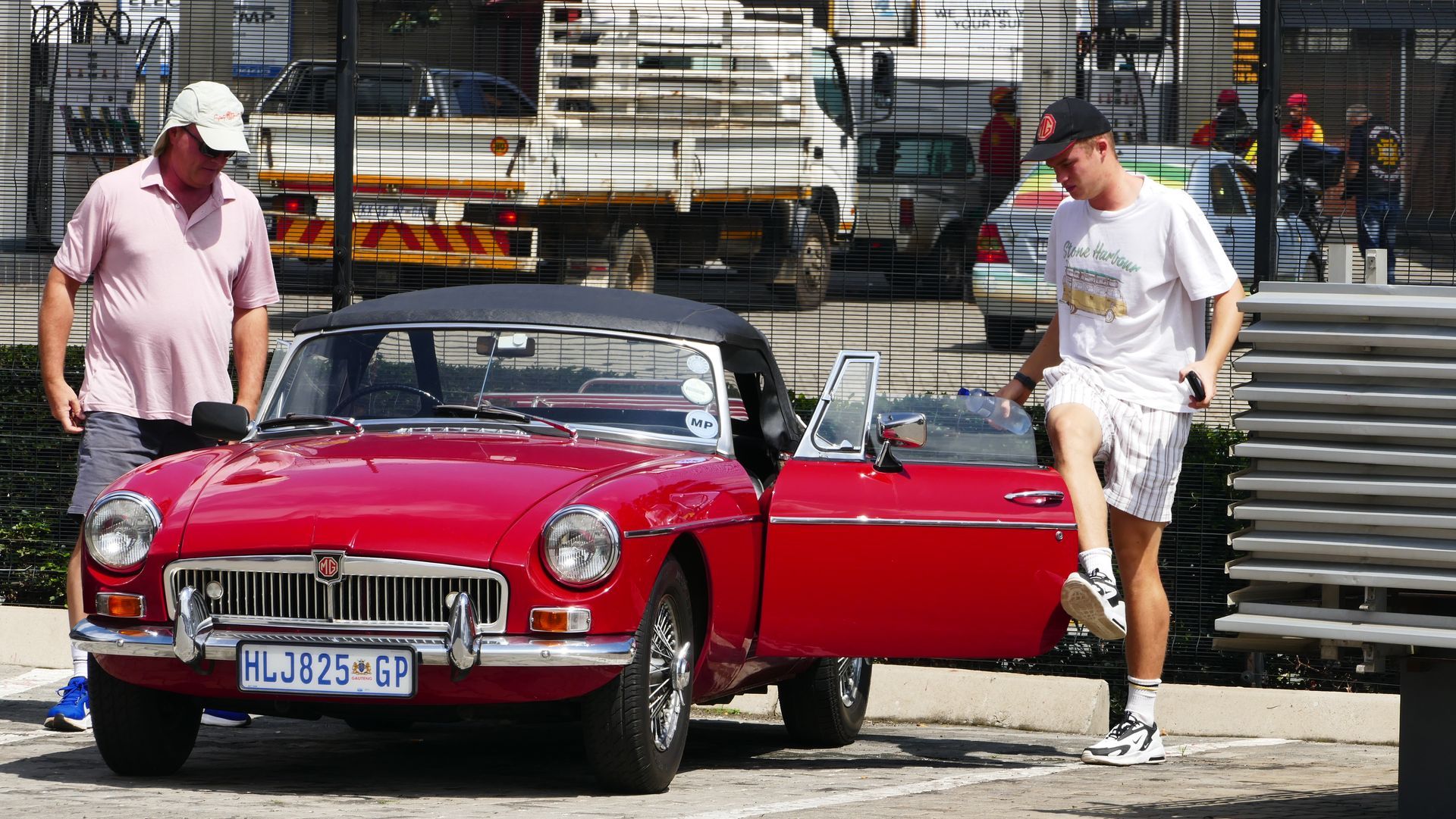 Red convertible car; two men nearby, one opening door, sunny outdoor setting.