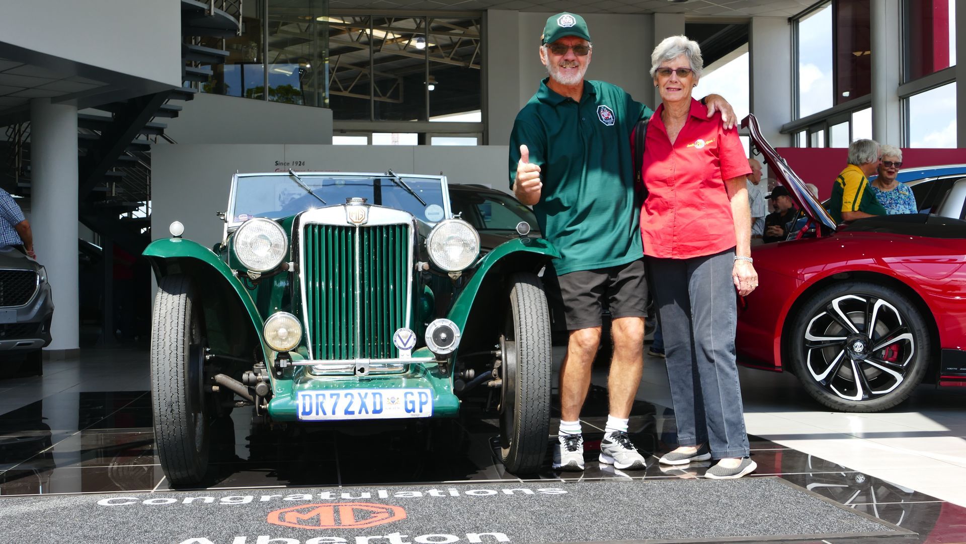 Couple poses with a classic green car; man gives thumbs up. Car in showroom.