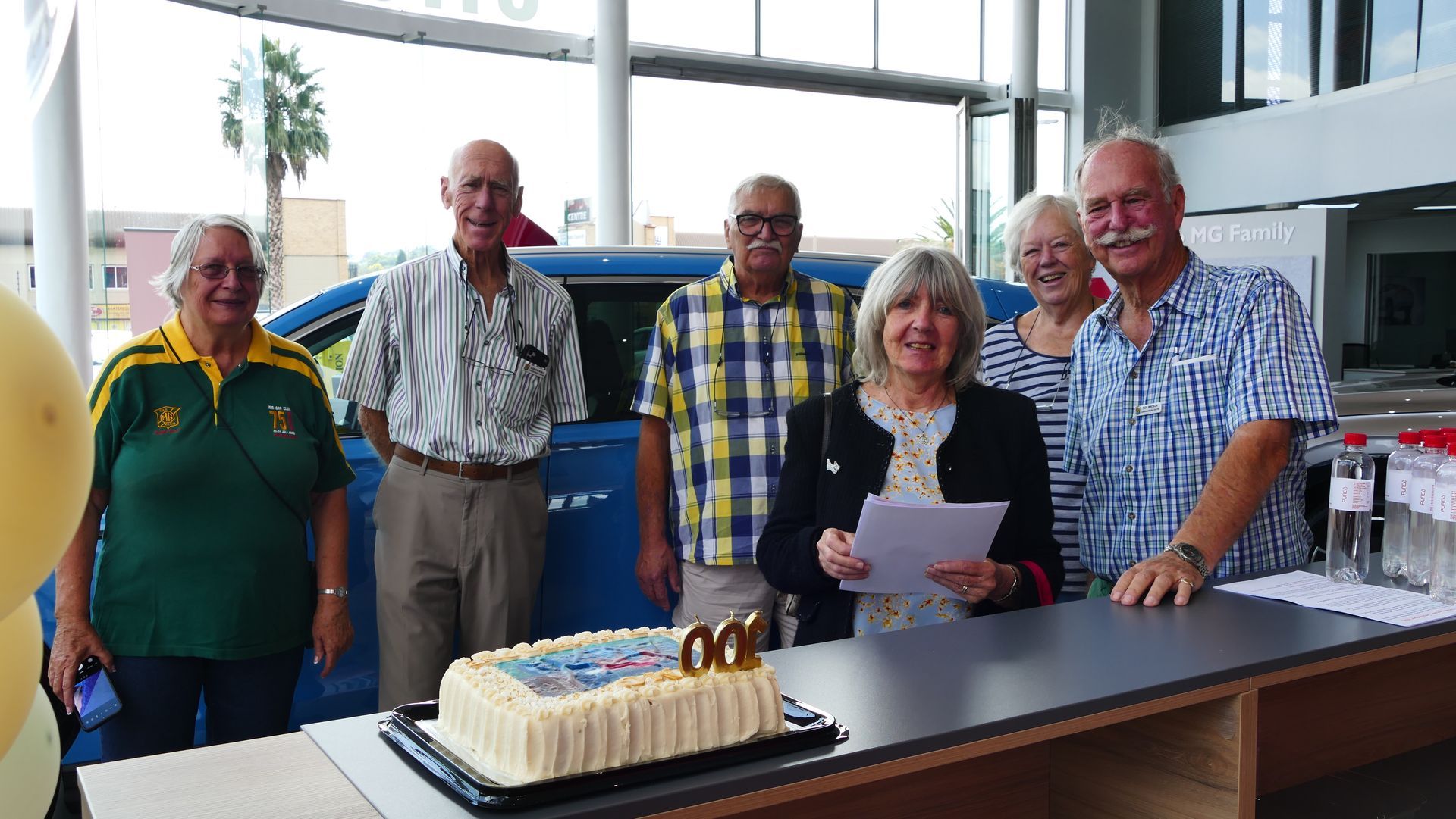 Group of people celebrate with cake in front of a blue car.  A woman holds paper, smiling.