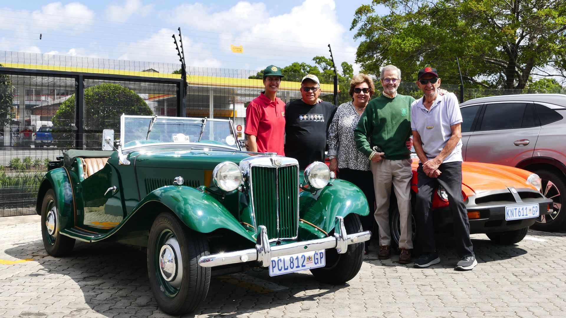 Group of people posing with green vintage car; sunny outdoor setting.