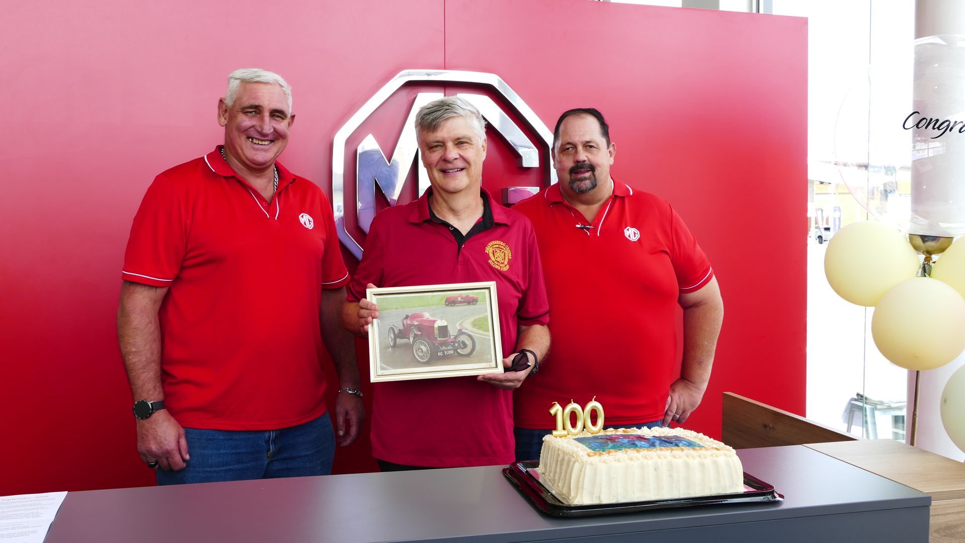 Three men in red shirts celebrate MG's 100th anniversary with a framed picture and cake in front of an MG logo.