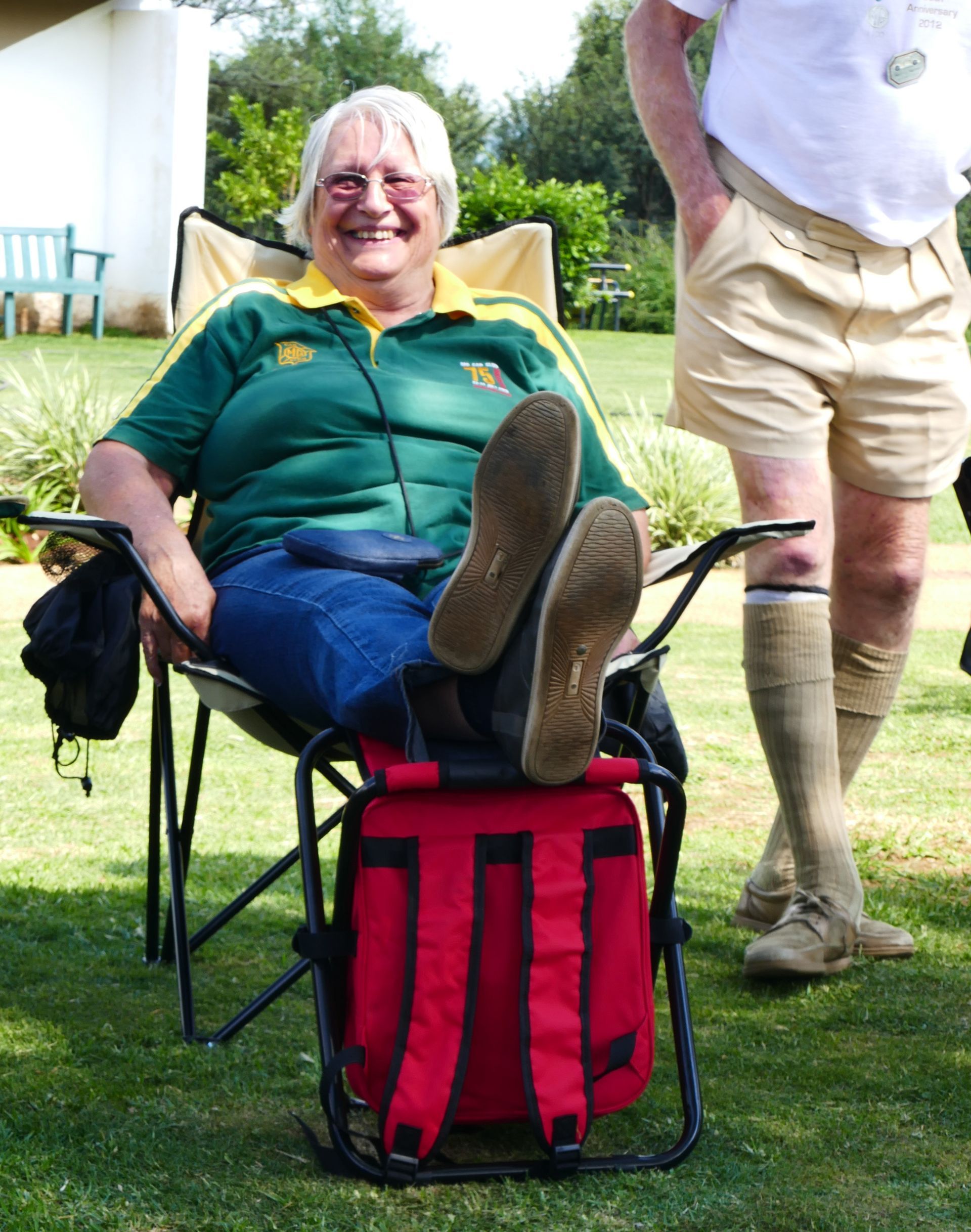 Woman in a folding chair, feet up on a red cooler, smiling in a grassy outdoor setting.