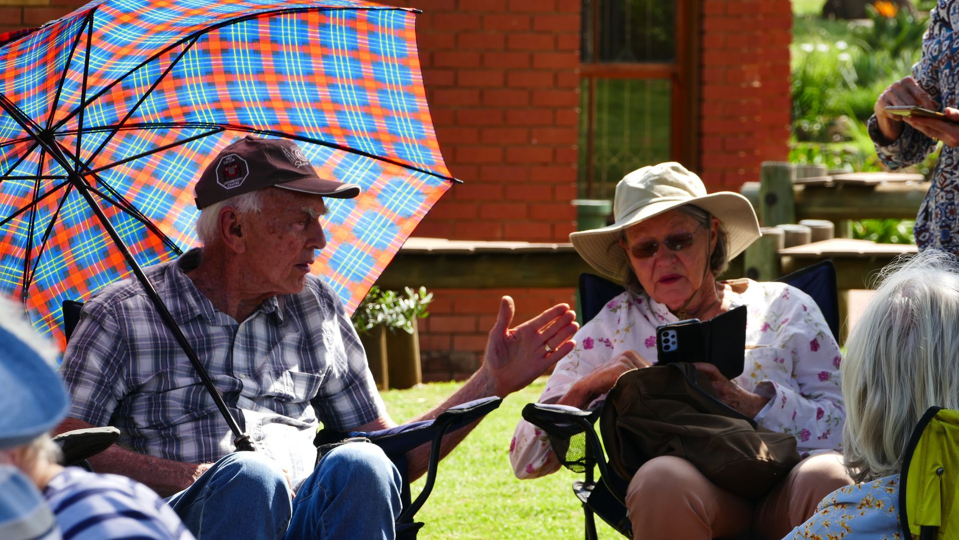 Two people outdoors in lawn chairs, shaded by umbrella. Man gestures while talking. Woman holds phone.