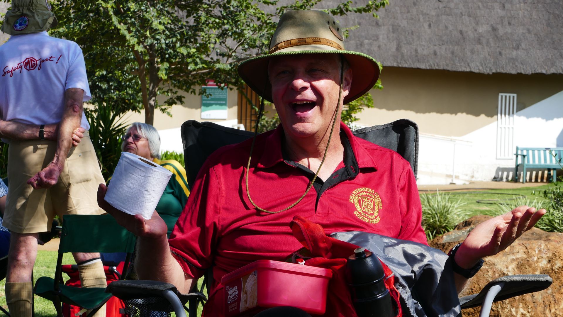 Man in a red shirt and hat seated outdoors, gesturing with open hands and smiling.