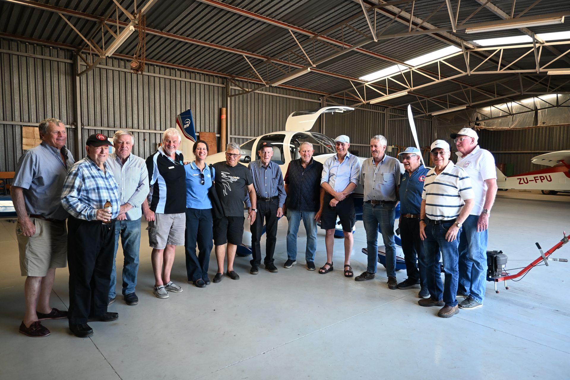 Group of people standing in a hangar with two small airplanes. Sunlight streams in.