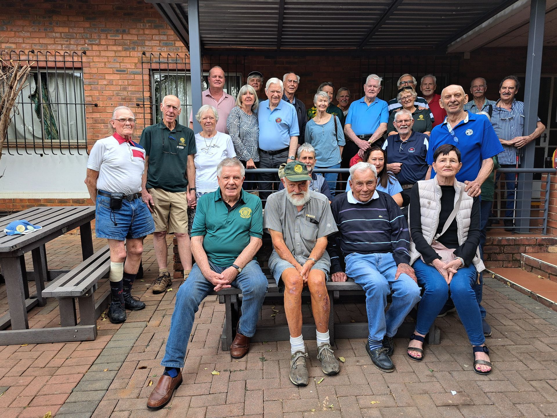 Group of people posing outdoors, brick building backdrop. Some seated on bench, others standing.