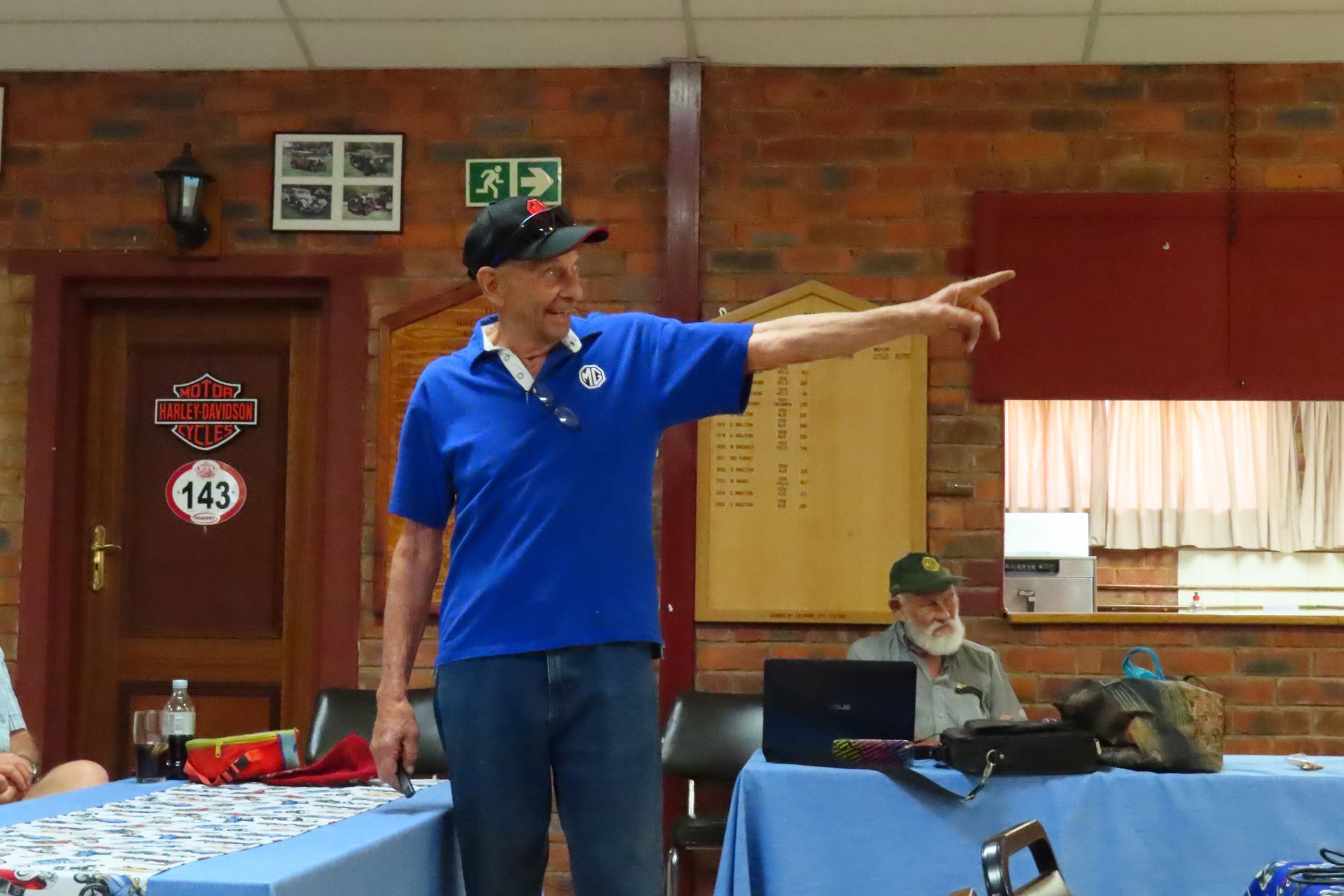 Man in blue shirt points, speaking at a table. Another man sits at a table with a laptop. Brick wall background.