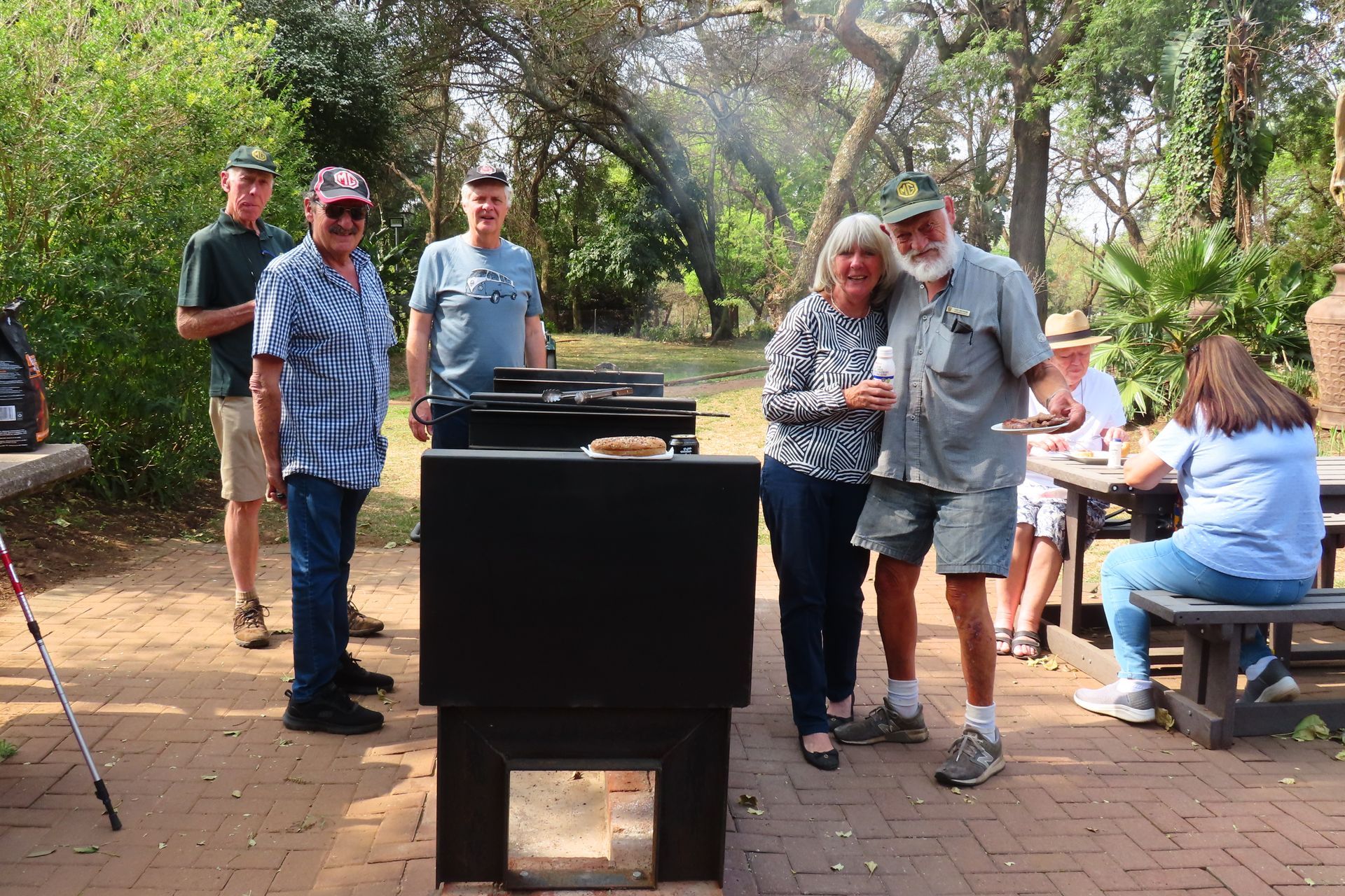 Group of people around a barbecue in a park, enjoying food and company.