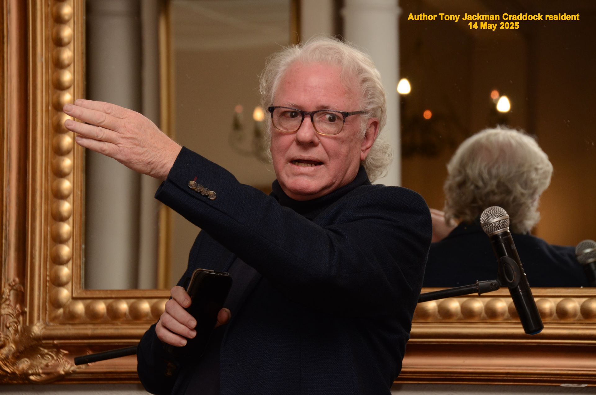 Man with glasses gestures while speaking at an event, with microphone and ornate mirror in the background.