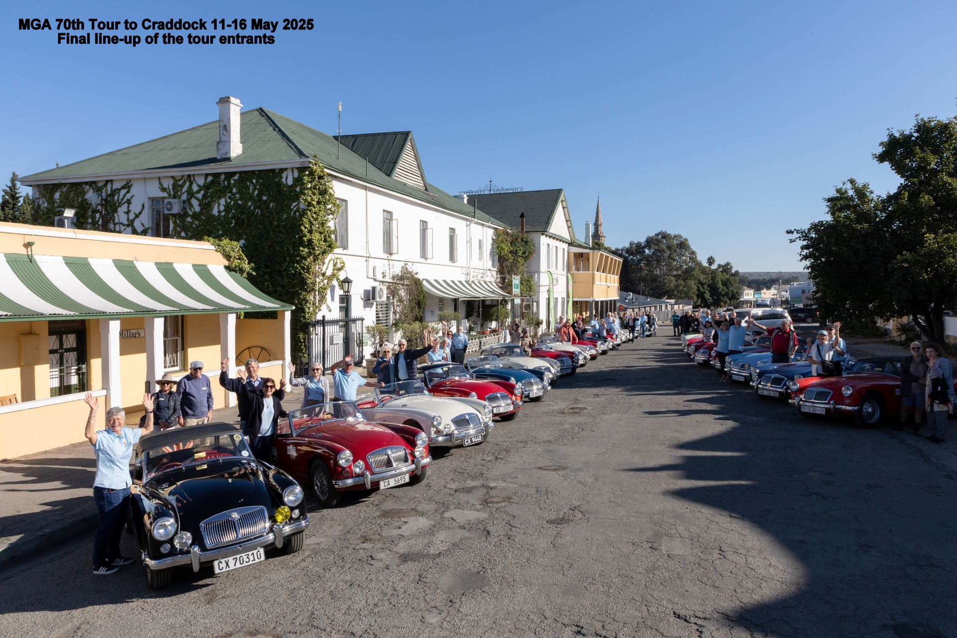 MGA cars line a street; people wave. Buildings with awnings and trees in background.