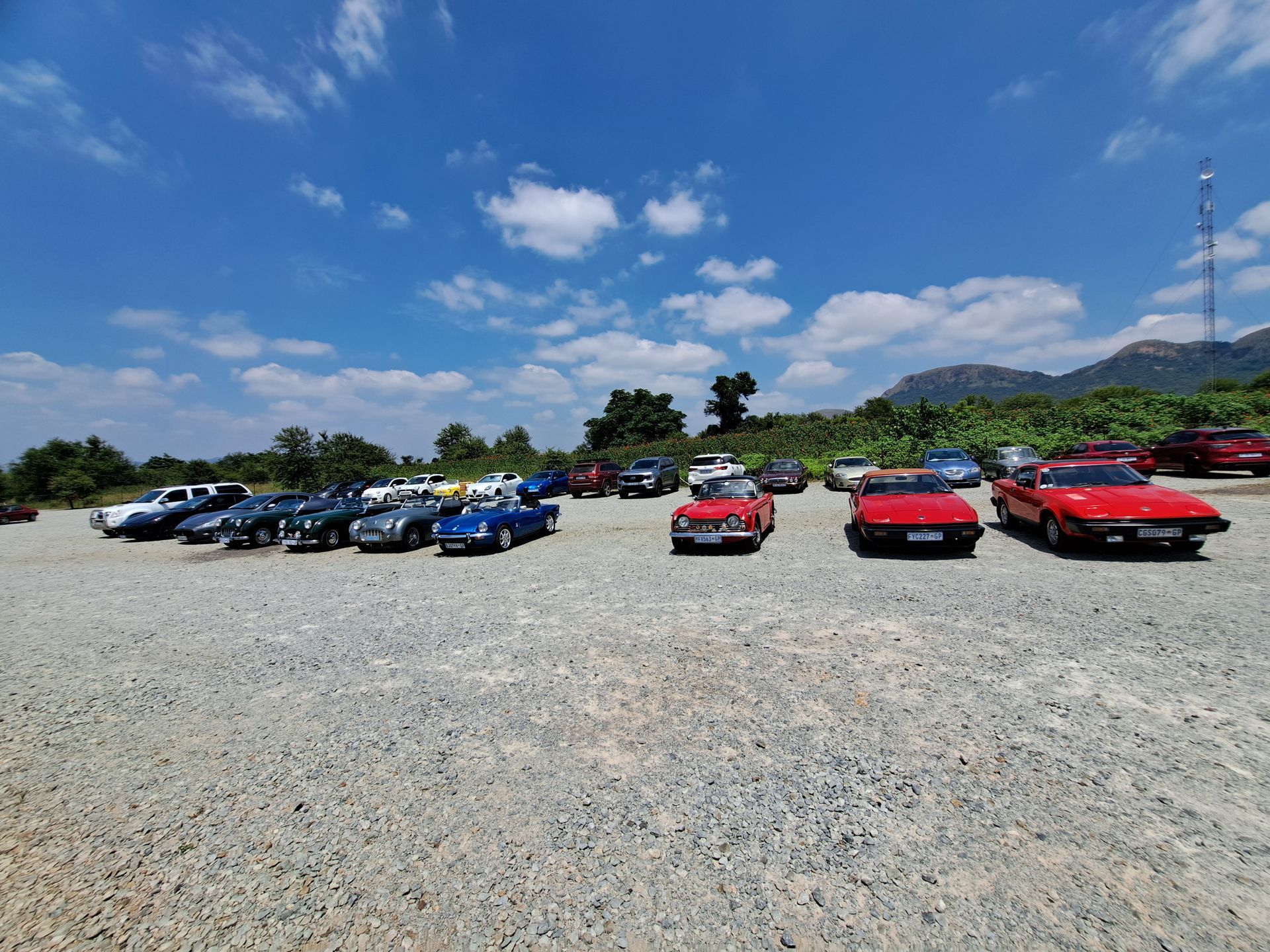 Classic cars parked in a gravel lot on a sunny day. Blue sky with clouds, trees, and mountains in background.