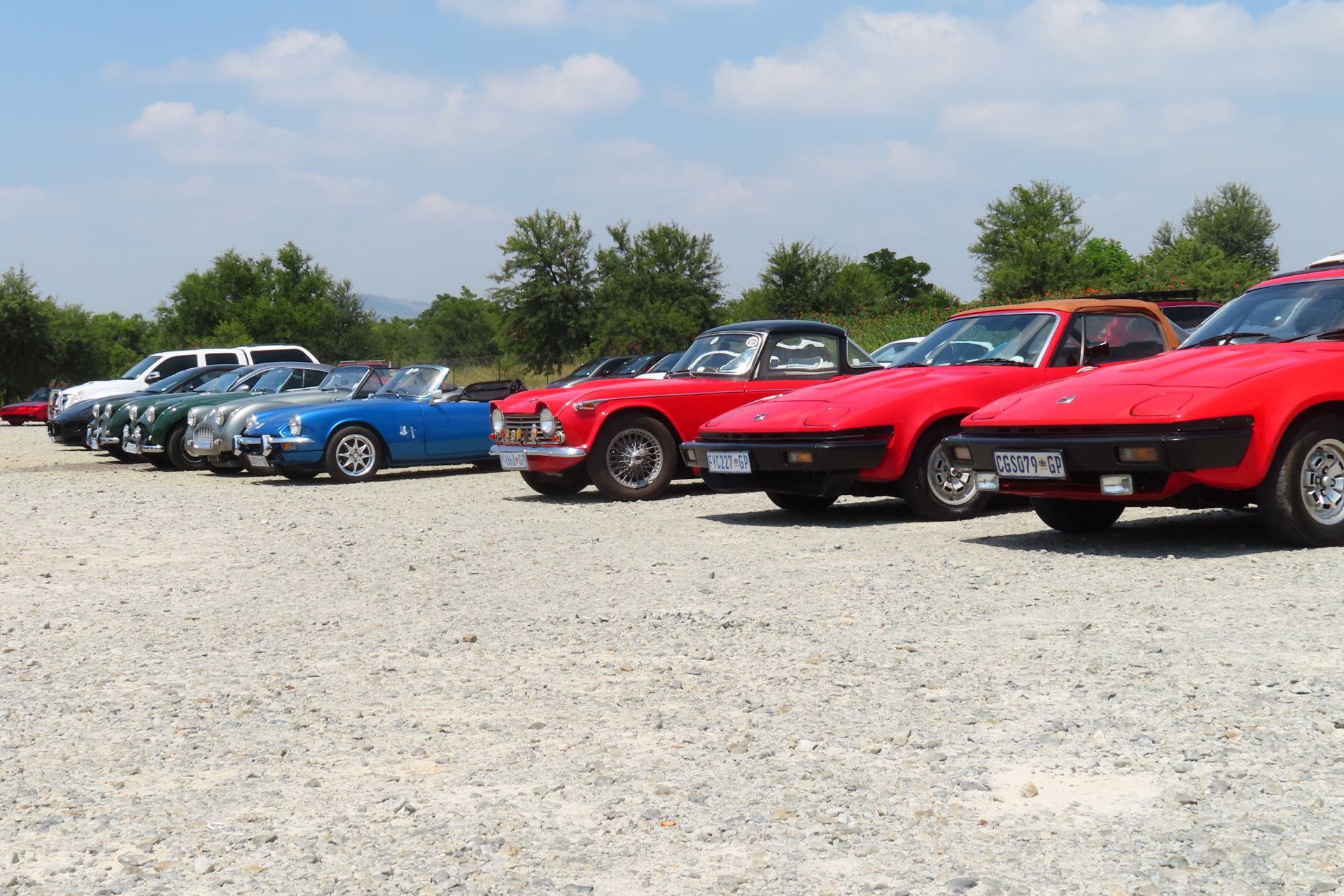 Classic red, blue, and green sports cars parked in a gravel lot on a sunny day.