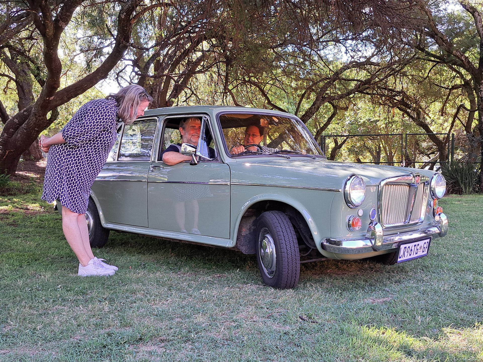 Woman leans towards a vintage green car with two people inside, in a grassy area with trees.