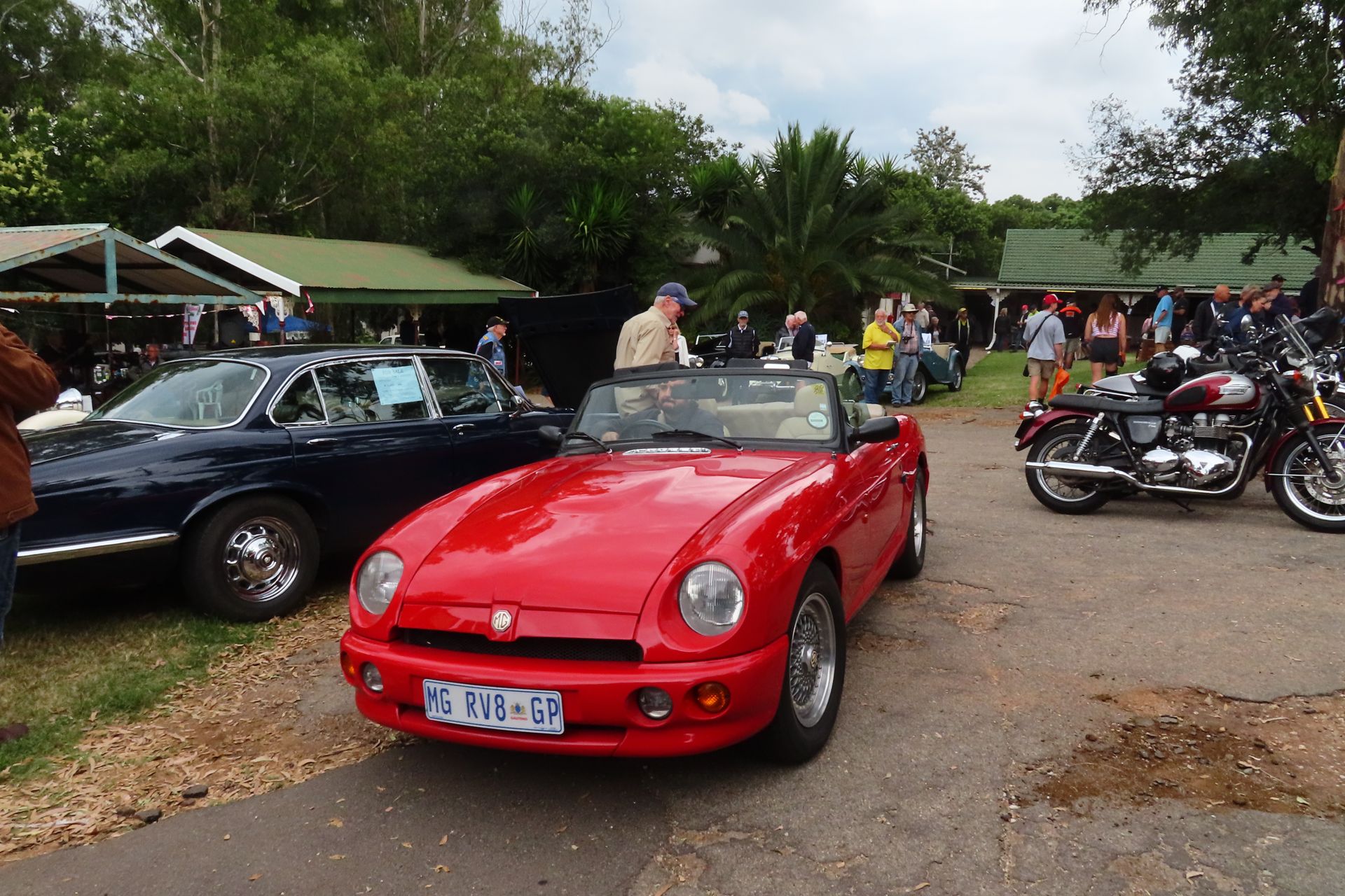 Red convertible and blue car parked at an outdoor event with motorcycles and people.