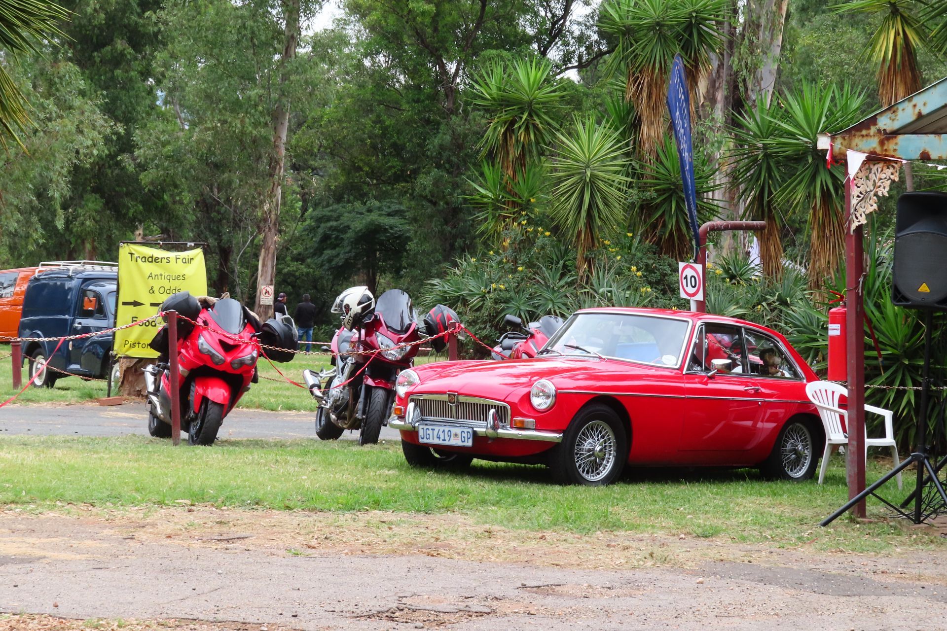 Red vintage car and motorcycles parked on grass near trees, a speaker, and a sign.