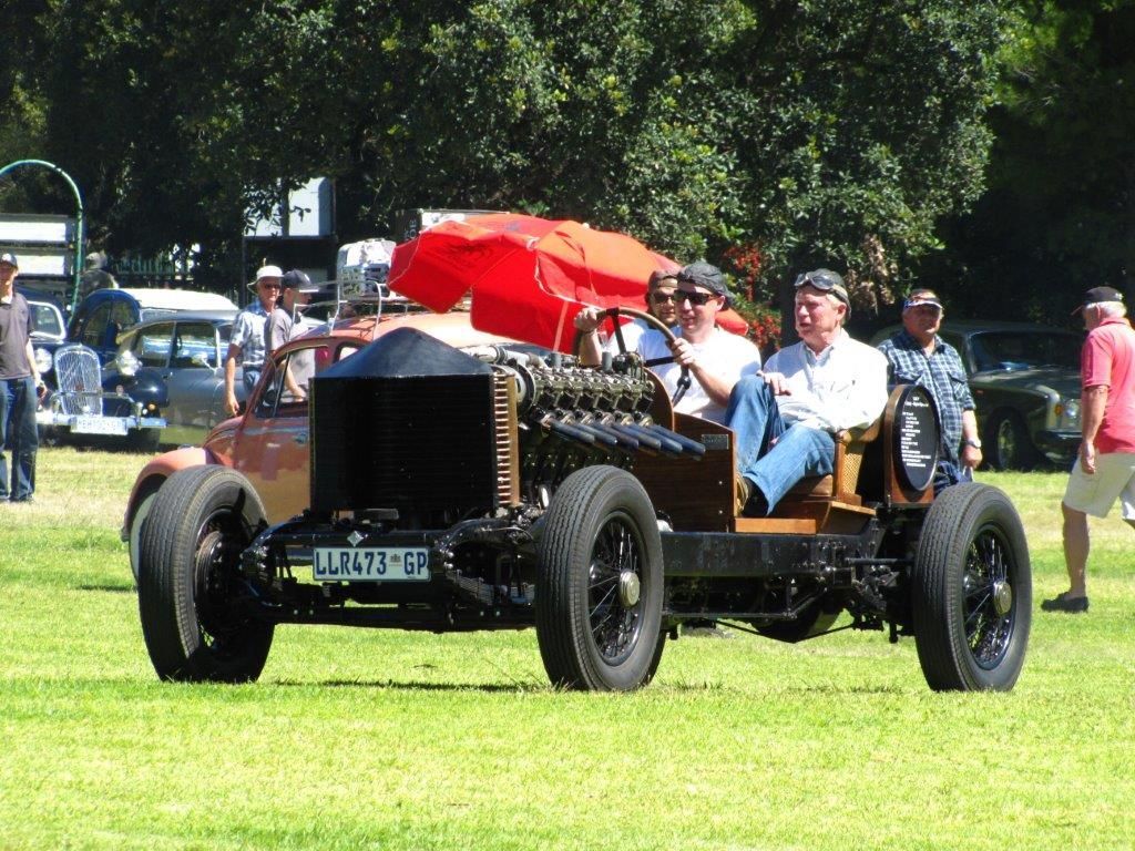 Vintage open-wheel car on grass, driven by two people. Red umbrella visible. Other vehicles and people in background.