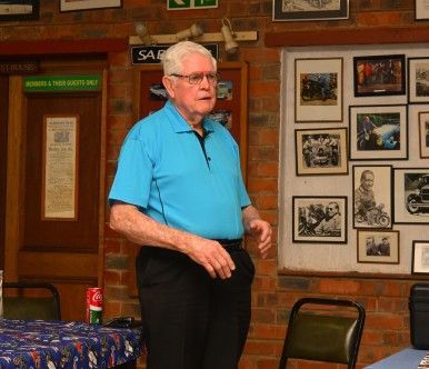 Man in a blue shirt and black pants speaks indoors, next to a wall with framed photos.