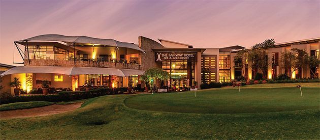 A golf course clubhouse at dusk, with lights on. Green lawn, buildings with modern architecture.