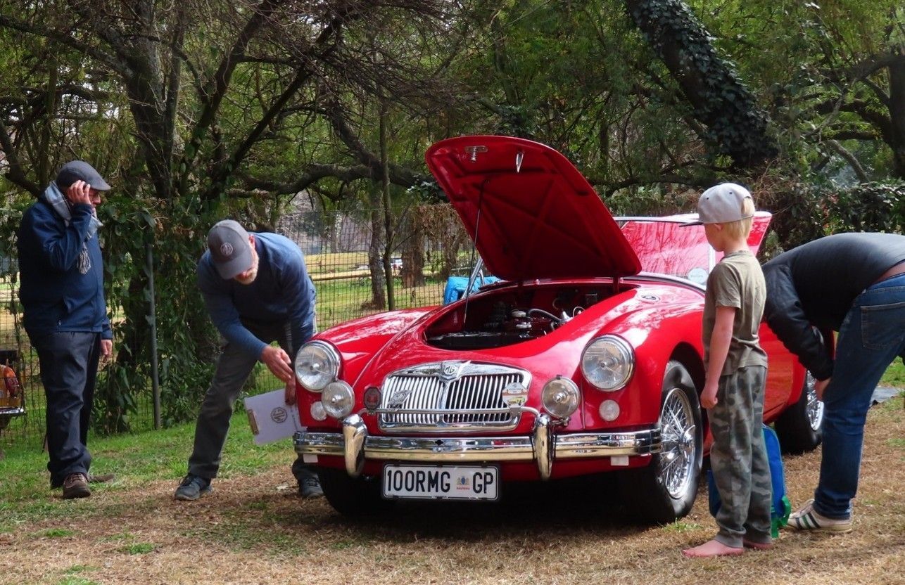 People examining a red vintage sports car with the hood open, outdoors.