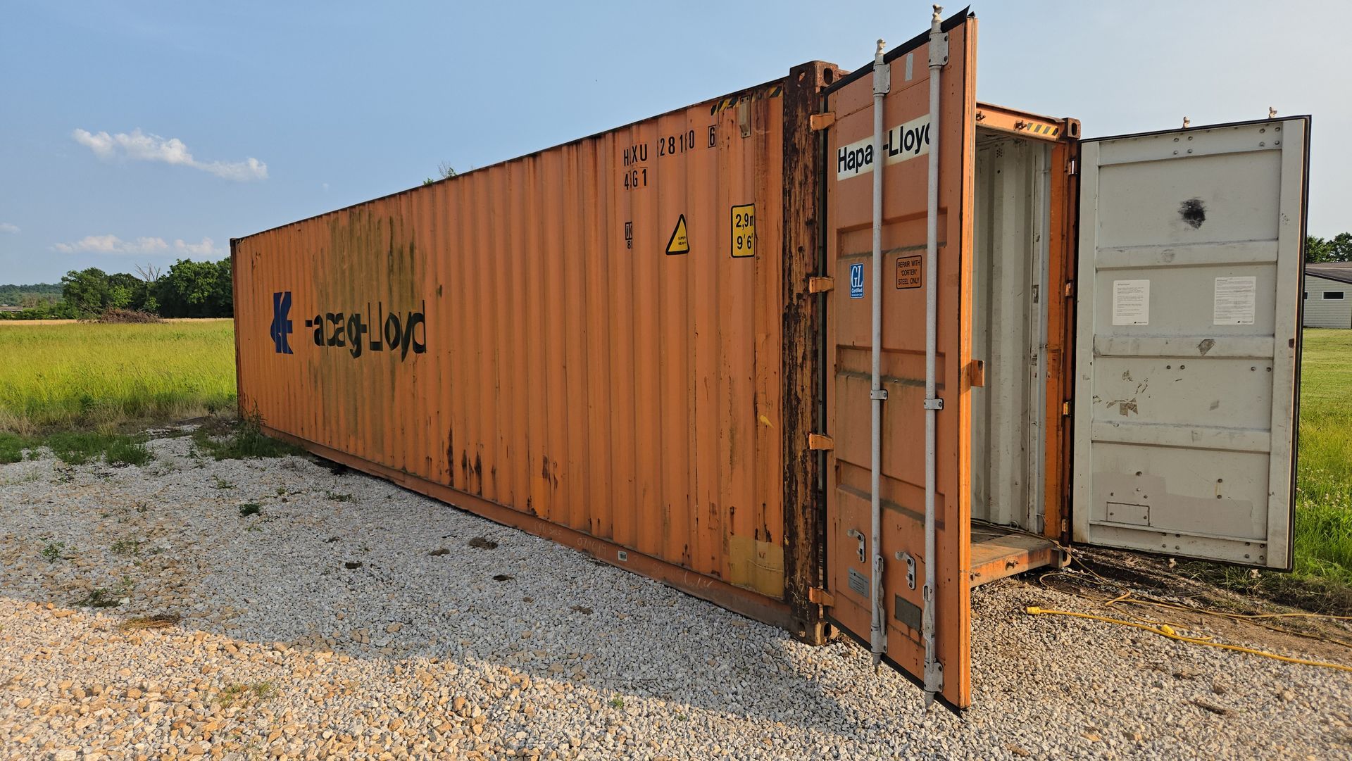Orange shipping container with open doors on gravel, in a field under a blue sky.