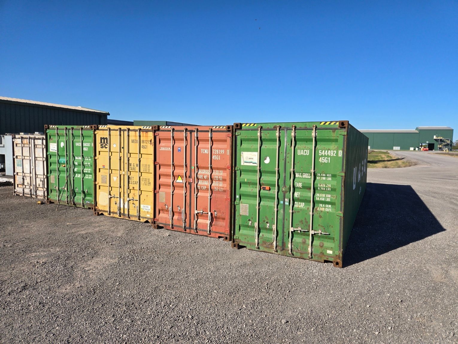 Line of colorful shipping containers on a gravel lot under a blue sky.