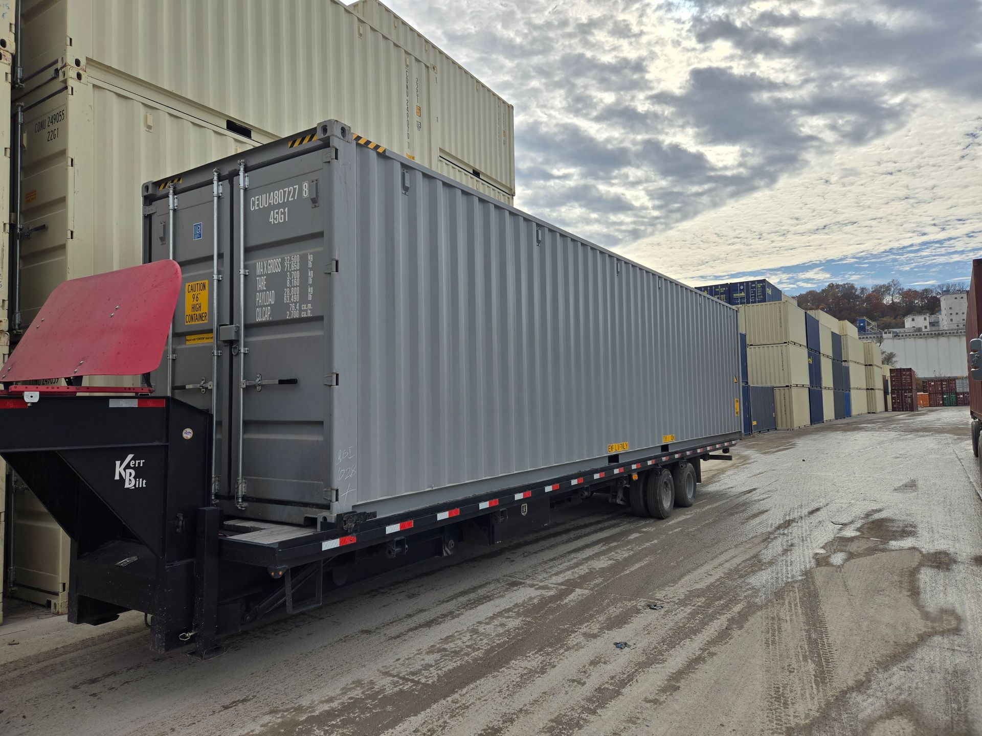 A gray shipping container on a trailer in a lot with other containers. Cloudy sky.