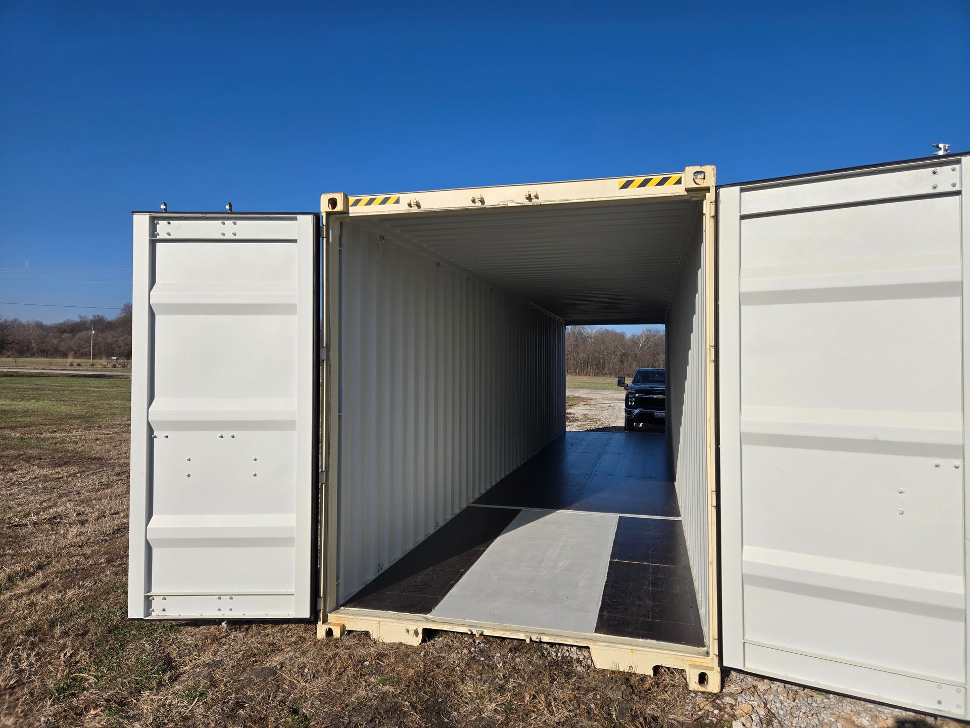 An open, empty cream-colored shipping container with doors ajar, on dirt ground, a vehicle visible in the distance.