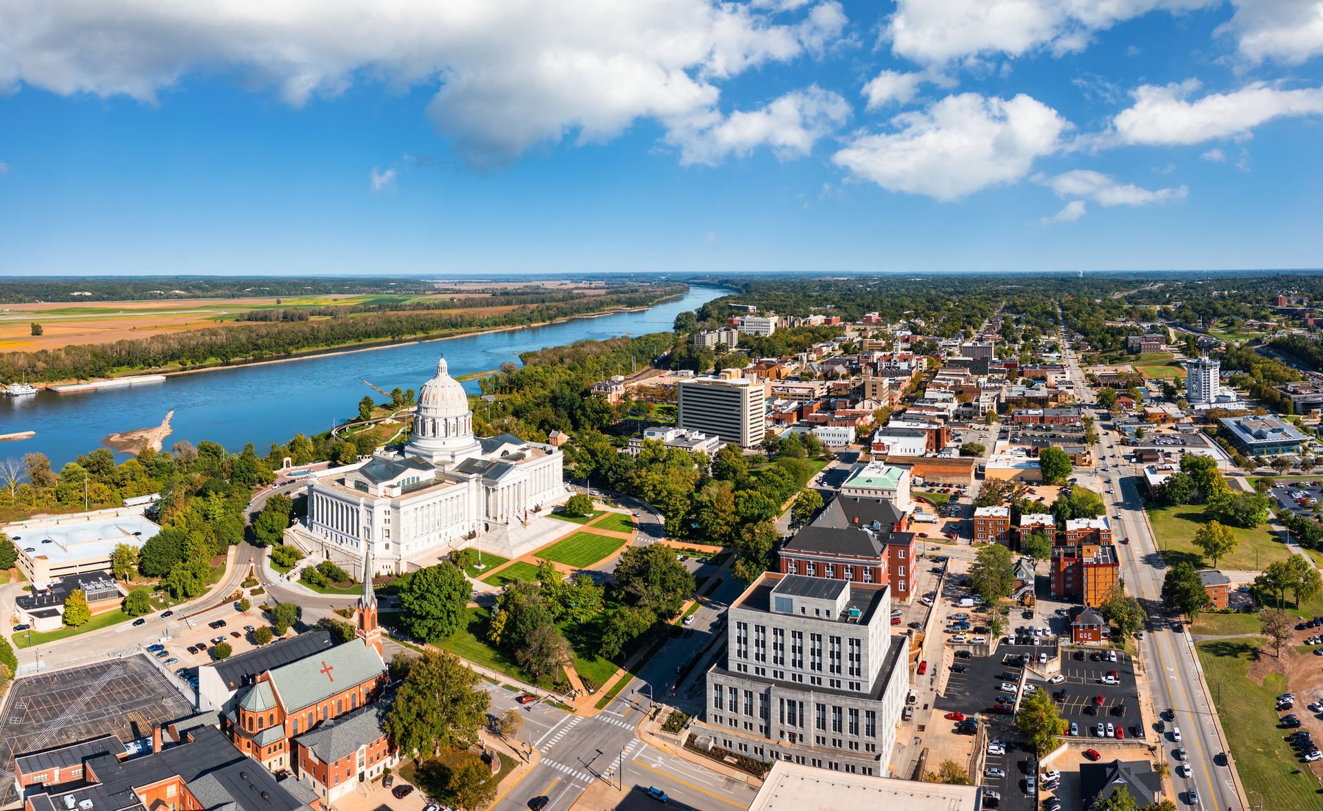 Aerial view of Jefferson City, Missouri, with the state capitol building near the Missouri River under a blue sky.