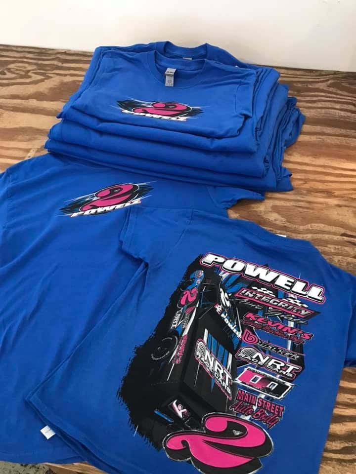 Blue t-shirts with racing logo stacked on a wooden surface. The shirts feature a design of a race car.