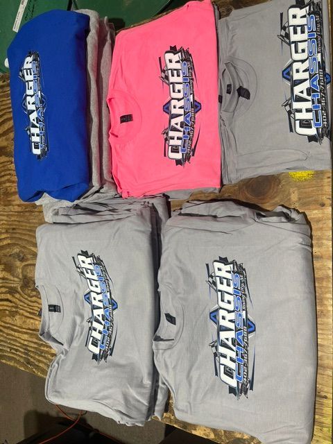 Five folded t-shirts, including one blue, one pink, and three grey, all printed with a Charger logo, stacked on a wood table.