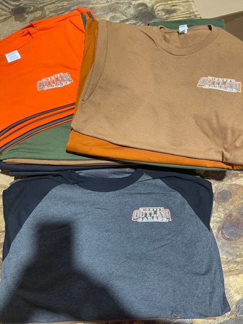A stack of folded t-shirts in various colors including orange, olive green, tan, and a gray baseball-style shirt.