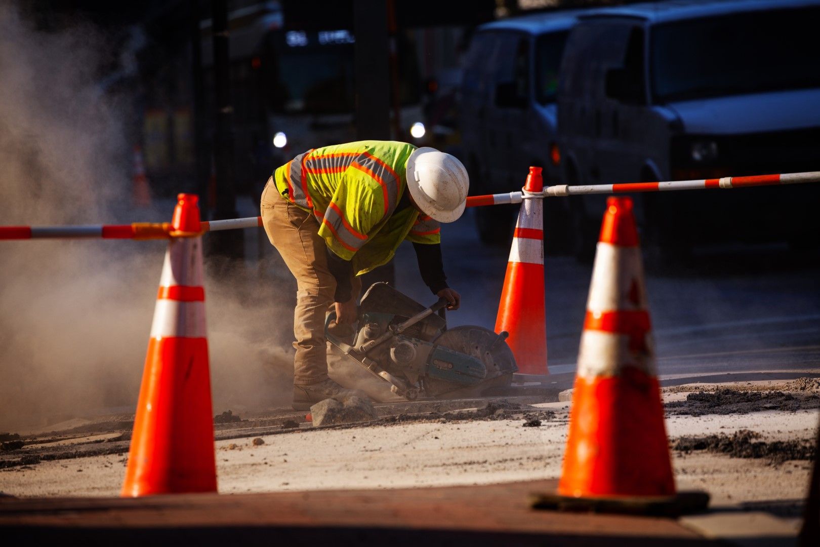 Construction worker in yellow vest and hard hat breaking up pavement with a jackhammer, surrounded by orange traffic cones.