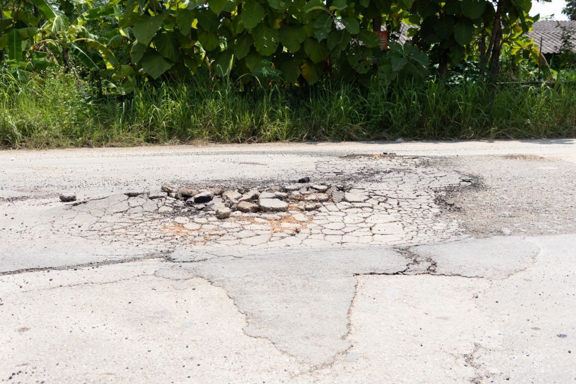 A cracked and crumbling asphalt road surface, highlighting a large pothole, with greenery in the background.