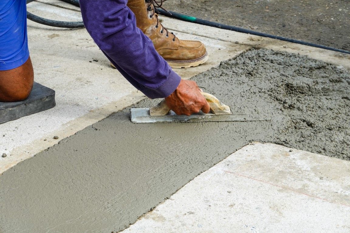 Person using a trowel to smooth wet concrete on a concrete surface outdoors. They wear a purple long-sleeved shirt and work boots.